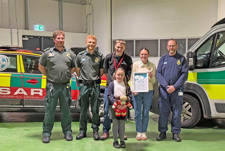 A child stands in an ambulance bay with her mother and four male emergency services professionals - two uniformed ambulance workers, a doctor from a medical charity, and an ambulance support staff member – after receiving a bravery award